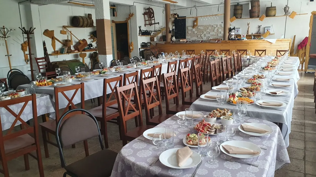Svinību zāle ar gariem klātiem galdiem un koka krēsliem, pie sienām senlietas