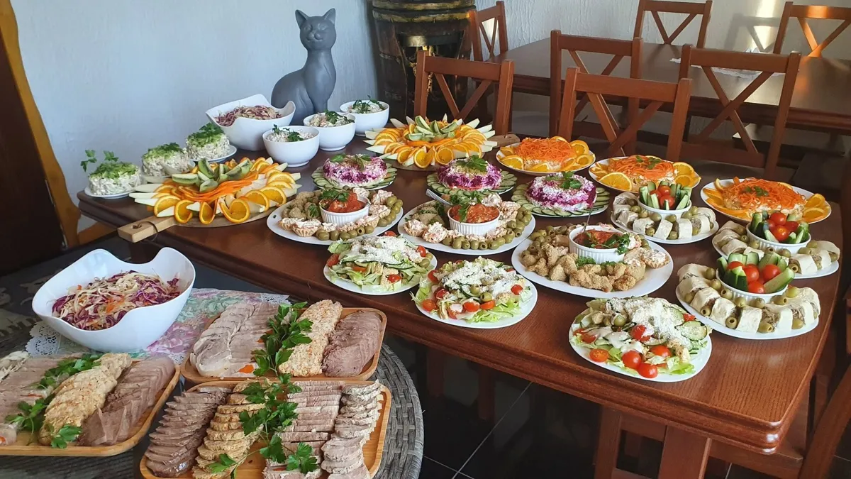 Galds ar salātiem, uzkodām un gaļas šķīvjiem, fonā pelēka kaķa figūra